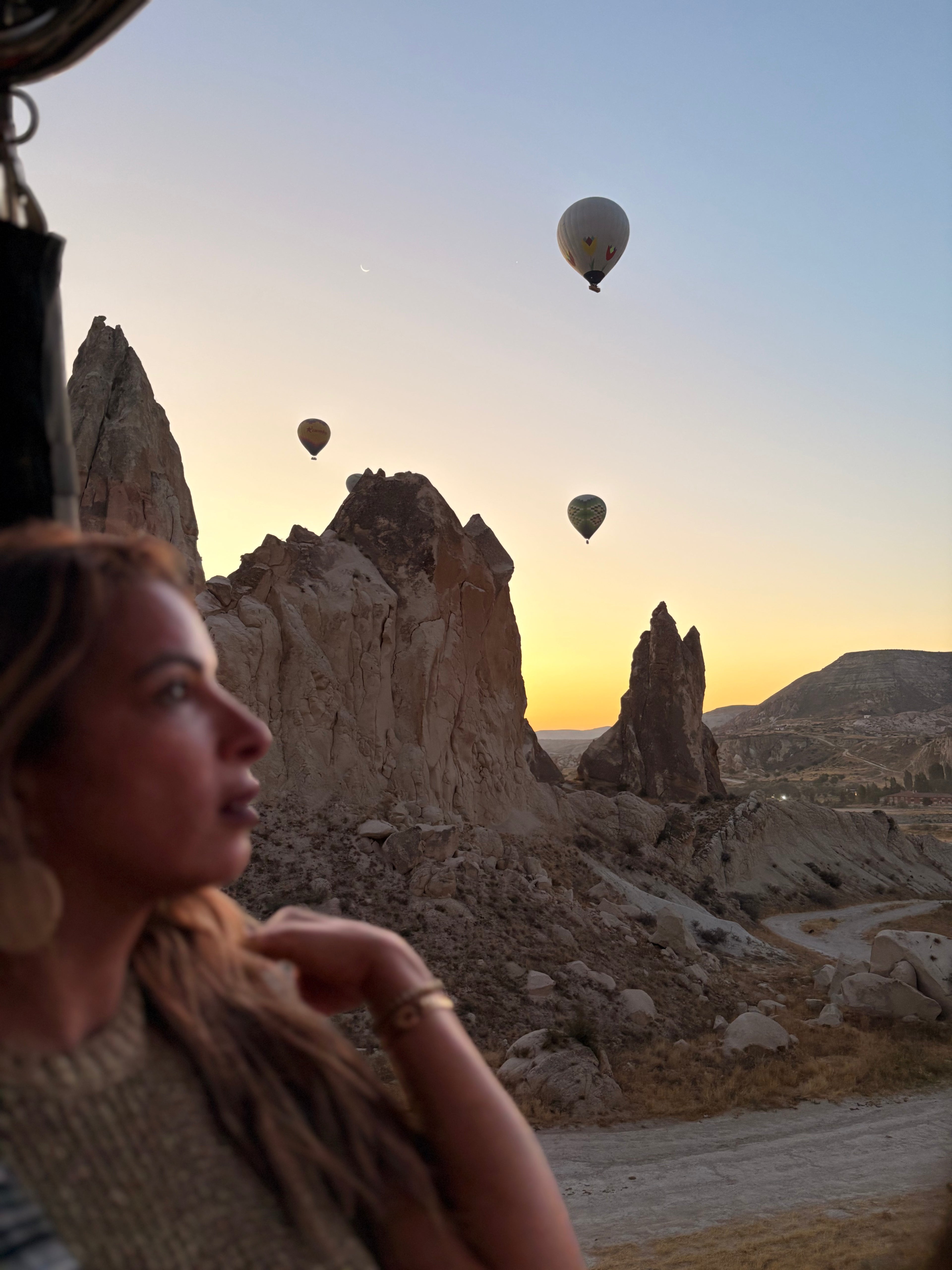 Woman looking at hot air balloons in a desert landscape during sunset.